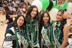 Four smiling senior girls wearing sashes and leis at a school event.