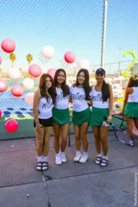 Four women in matching green skirts and white tops posing outdoors with colorful balloons.
