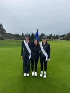 Three women in golf attire posing on a green field with sashes and flags.