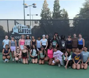 A diverse group of young people posing on a tennis court with a net and trees in the background.